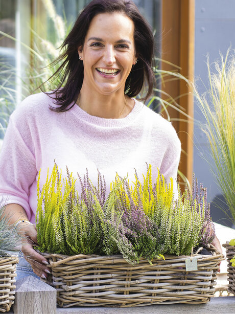 Blumen-Risse, Aktuelles, Trendthemen, Heide-macht-Laune, Frau-steht-auf-Veranda-und-haelt-Balkonkasten-mit-Heidebepflanzung