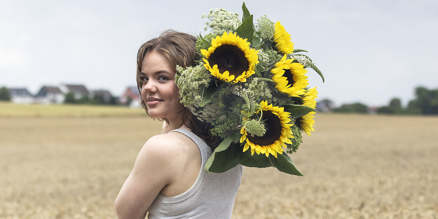 Blumen-Risse, Aktuelles, Aktion-Sonnenblumen, Frau-mit-Sonnenblumen-ueber-Schulter-steht-im-Feld