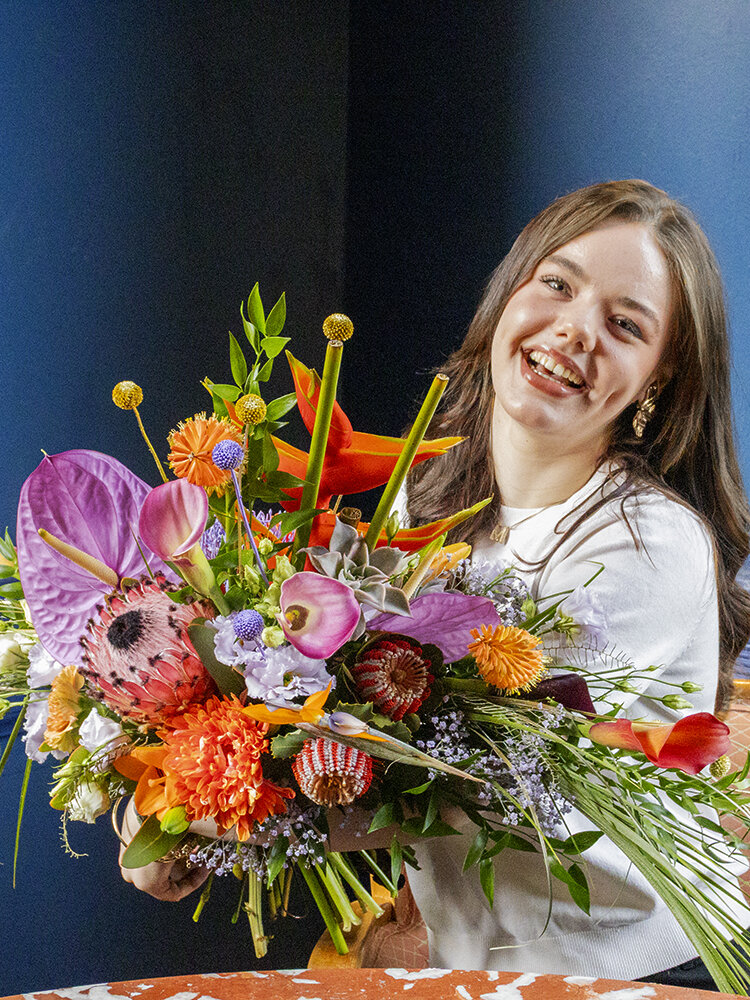 Junge Frau sitzt mit opulentem Blumenstrauß an Tisch vor blauer Wand