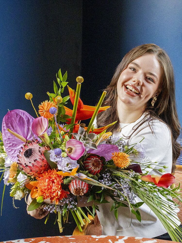 Junge Frau sitzt mit opulentem Blumenstrauß an Tisch vor blauer Wand