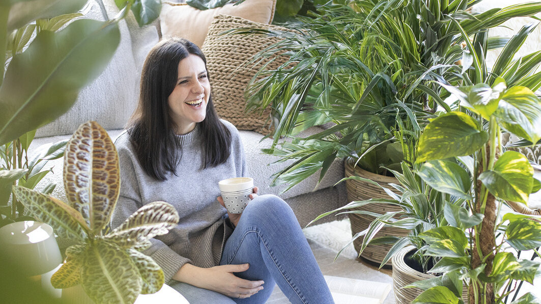 Blumen-Risse, Zimmerpflanzen, Gruenpflanzen, Titelbild, Frau-mit-Kaffeetasse-sitzt-im-Wohnzimmer-zwischen-Gruenpflanzen-auf-Boden