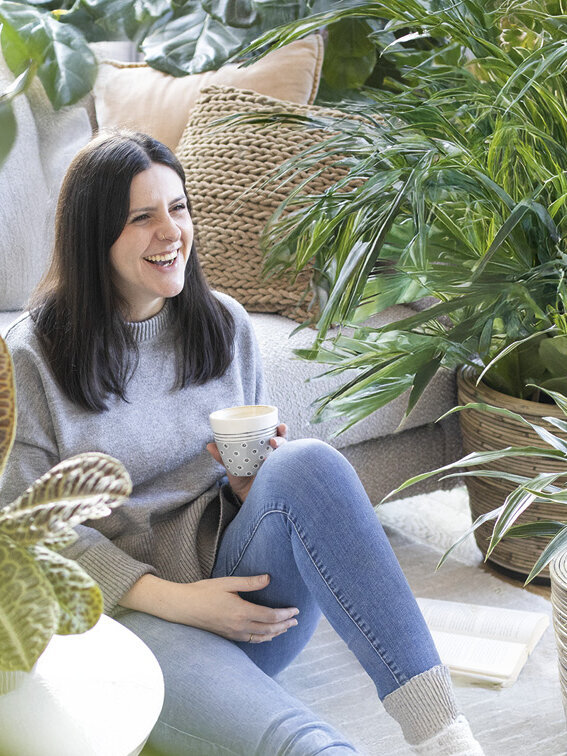 Blumen-Risse, Zimmerpflanzen, Gruenpflanzen, Titelbild, Frau-mit-Kaffeetasse-sitzt-im-Wohnzimmer-zwischen-Gruenpflanzen-auf-Boden