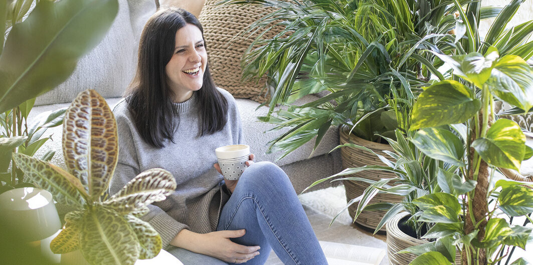 Blumen-Risse, Zimmerpflanzen, Gruenpflanzen, Titelbild, Frau-mit-Kaffeetasse-sitzt-im-Wohnzimmer-zwischen-Gruenpflanzen-auf-Boden