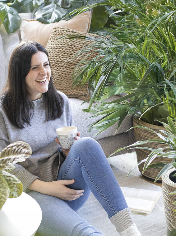 Blumen-Risse, Zimmerpflanzen, Gruenpflanzen, Titelbild, Frau-mit-Kaffeetasse-sitzt-im-Wohnzimmer-zwischen-Gruenpflanzen-auf-Boden