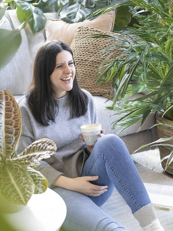 Blumen-Risse, Zimmerpflanzen, Gruenpflanzen, Titelbild, Frau-mit-Kaffeetasse-sitzt-im-Wohnzimmer-zwischen-Gruenpflanzen-auf-Boden