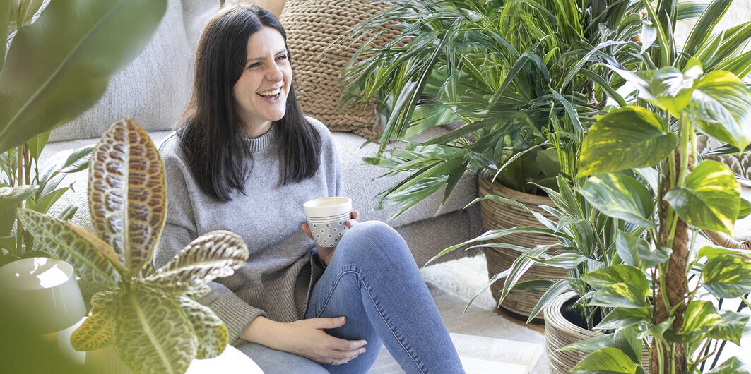 Blumen-Risse, Zimmerpflanzen, Gruenpflanzen, Titelbild, Frau-mit-Kaffeetasse-sitzt-im-Wohnzimmer-zwischen-Gruenpflanzen-auf-Boden