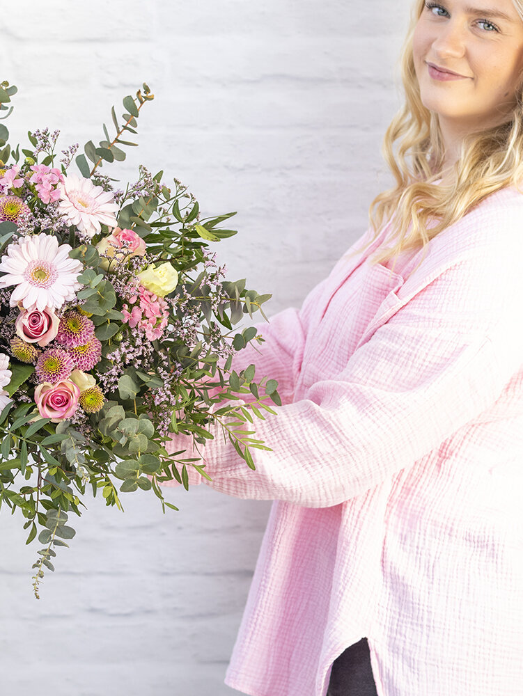 Blumen-Risse, Blumenversand, SW002-Hauptsache-Pink, Rosa, Pink, Frau-haelt-rosafarbenen-Blumenstrauss-mit-zwei-Haenden-vor-weisser-Steinwand