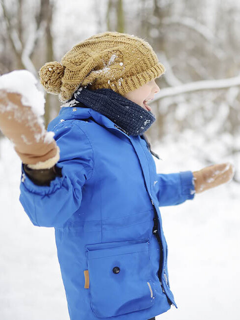 Winter im Garten, Kind mit blauer Jacke und Handschuhen im Wintergarten mit Schneeball in der Hand