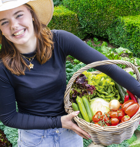 Blumen-Risse, Titelbild, Aktuelles, Selbstversorger, Frau-hält-vollen-Gemüsekorb-vor-Nutzbeet